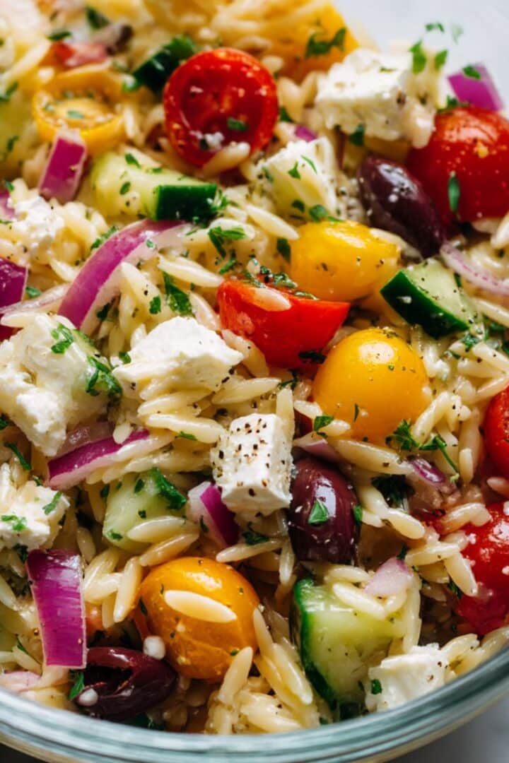 Overhead view of a vibrant Greek orzo salad filled with cherry tomatoes, cucumbers, black olives, red onions, feta cheese, and fresh parsley, all tossed in a light dressing and sprinkled with cracked black pepper.