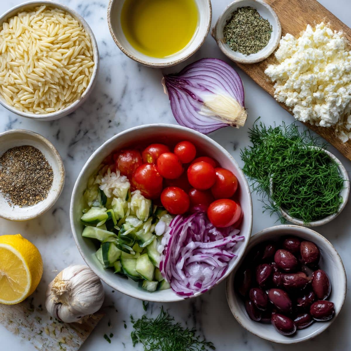 Overhead view of orzo pasta, tomatoes, cucumber, olives, feta, and herbs on a white counter. Fresh ingredients for homemade Greek orzo salad recipe.