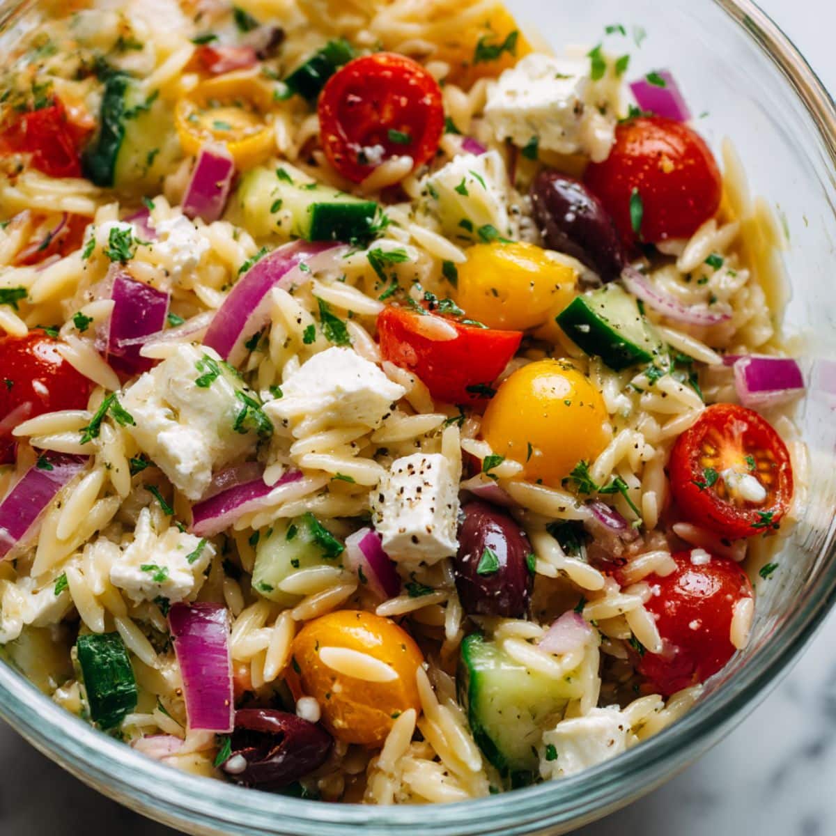 Overhead view of a vibrant Greek orzo salad filled with cherry tomatoes, cucumbers, black olives, red onions, feta cheese, and fresh parsley, all tossed in a light dressing and sprinkled with cracked black pepper.