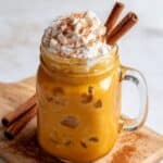 Iced pumpkin spice latte served in a mason jar, topped with whipped cream and cinnamon sticks, with rich golden tones and creamy foam spilling over the rim, styled on a rustic wooden board with cinnamon dust.