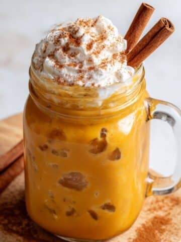 Iced pumpkin spice latte served in a mason jar, topped with whipped cream and cinnamon sticks, with rich golden tones and creamy foam spilling over the rim, styled on a rustic wooden board with cinnamon dust.