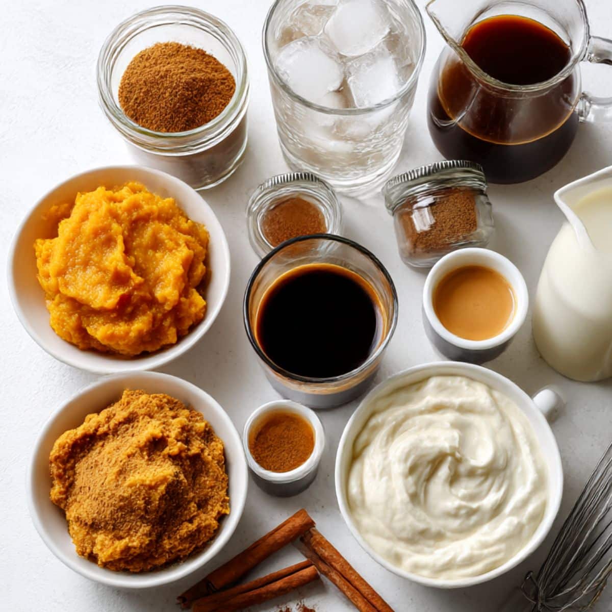 Realistic top-down image of iced pumpkin spice latte ingredients on a white kitchen counter with no labels, including pumpkin puree, spices, coffee, milk, and whipped cream tools in a cozy fall kitchen setting.