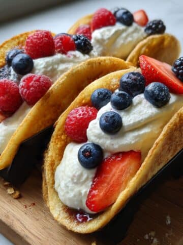 Pancake tacos filled with fresh berries and whipped cream in black taco holders on wooden serving board.