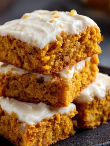 Moist pumpkin bars with cream cheese frosting stacked on a plate, showing rich orange crumb and smooth whipped topping.