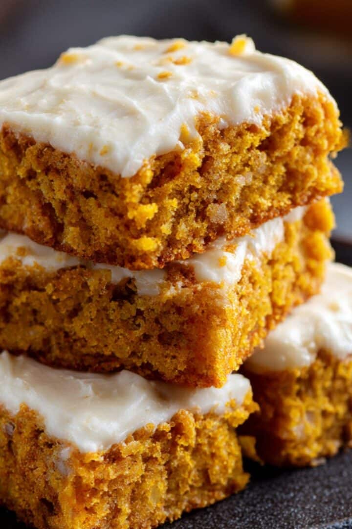 Moist pumpkin bars with cream cheese frosting stacked on a plate, showing rich orange crumb and smooth whipped topping.