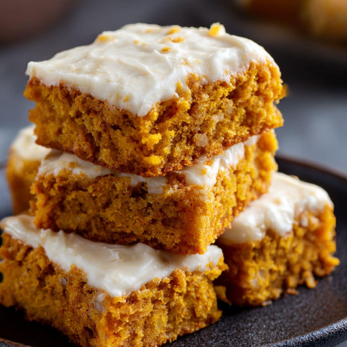 Moist pumpkin bars with cream cheese frosting stacked on a plate, showing rich orange crumb and smooth whipped topping.