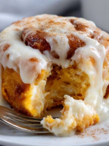 Moist pumpkin cinnamon roll with vibrant orange dough, topped with melted cream cheese frosting and gooey cinnamon filling on a white plate.