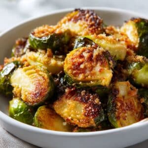 Crispy roasted Brussel sprouts with golden-brown panko crust, glistening with olive oil and topped with sea salt, served in a white bowl with vibrant green and caramelized edges.
