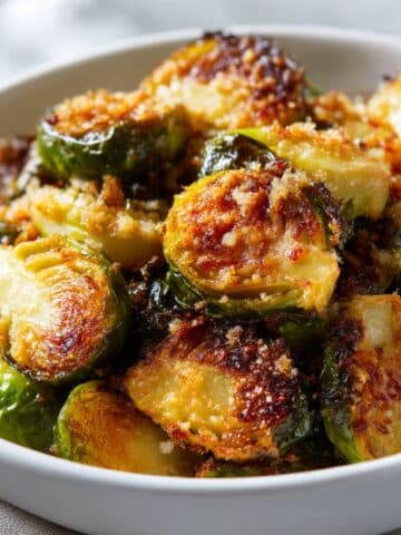 Crispy roasted Brussel sprouts with golden-brown panko crust, glistening with olive oil and topped with sea salt, served in a white bowl with vibrant green and caramelized edges.