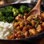 Glossy sesame chicken coated in a rich, sticky soy-garlic glaze, garnished with sesame seeds and sliced green onions, served in a black skillet alongside fluffy white rice and steamed broccoli.