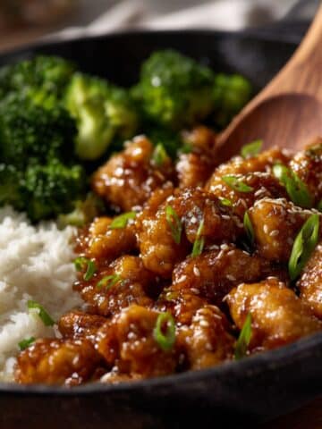 Glossy sesame chicken coated in a rich, sticky soy-garlic glaze, garnished with sesame seeds and sliced green onions, served in a black skillet alongside fluffy white rice and steamed broccoli.