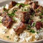 Close-up of juicy steak and queso rice with tender seared steak cubes smothered in creamy beige cheese sauce, served over fluffy white rice and topped with chopped fresh parsley.