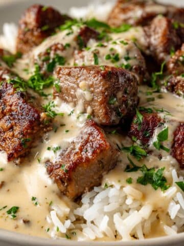 Close-up of juicy steak and queso rice with tender seared steak cubes smothered in creamy beige cheese sauce, served over fluffy white rice and topped with chopped fresh parsley.