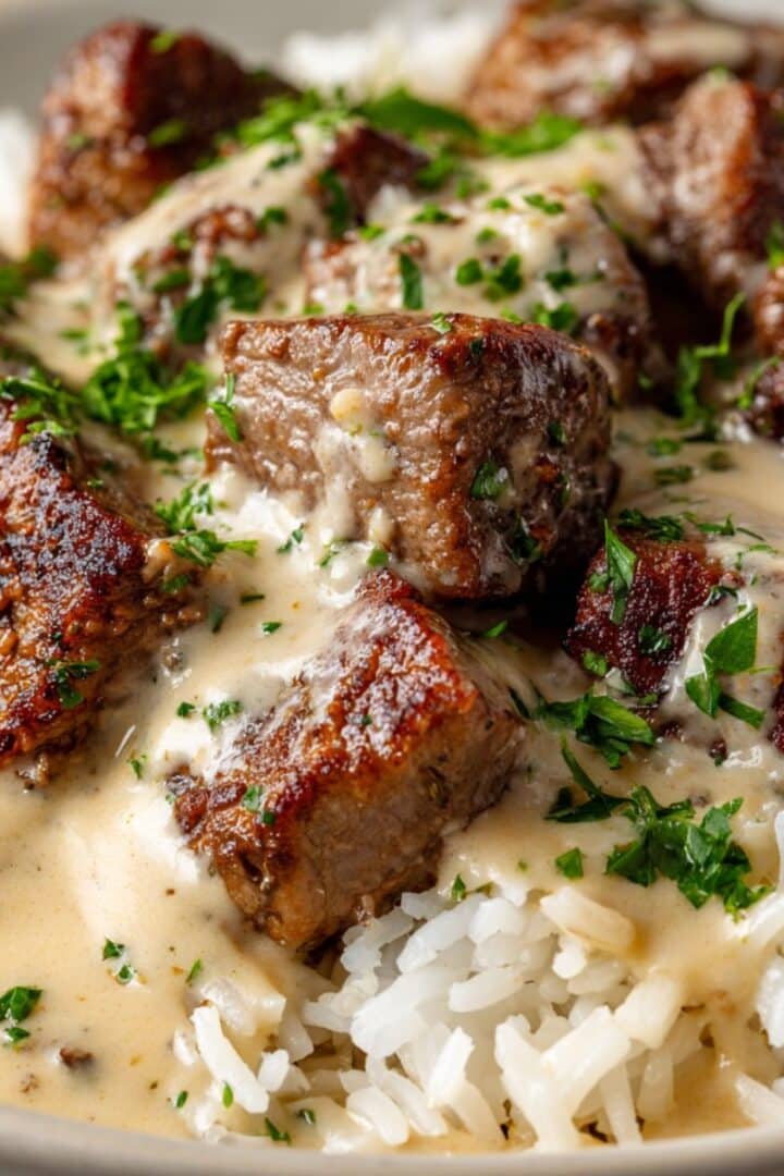 Close-up of juicy steak and queso rice with tender seared steak cubes smothered in creamy beige cheese sauce, served over fluffy white rice and topped with chopped fresh parsley.