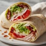 Turkey club wrap cut in half showing layers of deli turkey, bacon, lettuce and tomato in flour tortilla.