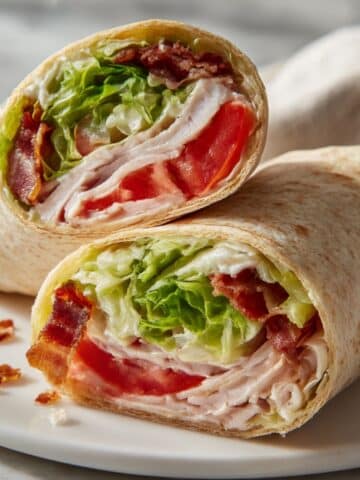 Turkey club wrap cut in half showing layers of deli turkey, bacon, lettuce and tomato in flour tortilla.