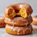 Stacked baked pumpkin donuts topped with glossy cinnamon glaze, with the top donut bitten to reveal a moist, fluffy orange interior. Rich fall colors and soft lighting highlight the glistening glaze and tender crumb on a clean white background.