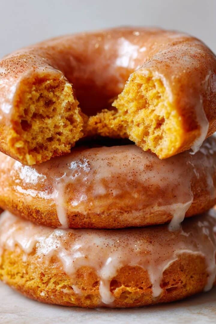 Stacked baked pumpkin donuts topped with glossy cinnamon glaze, with the top donut bitten to reveal a moist, fluffy orange interior. Rich fall colors and soft lighting highlight the glistening glaze and tender crumb on a clean white background.