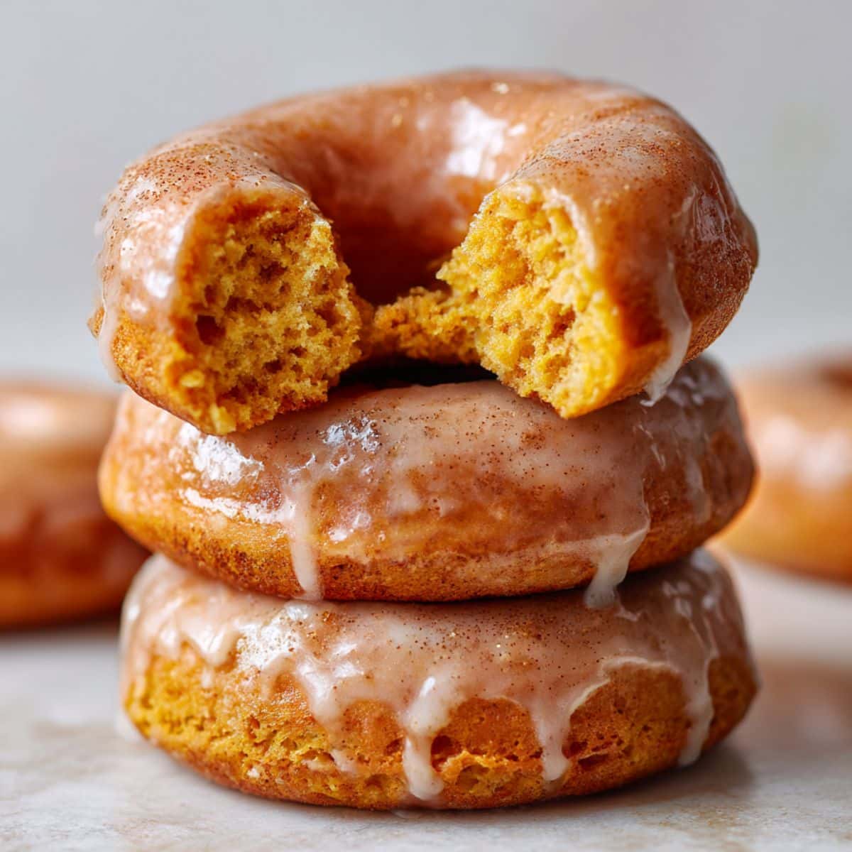 Stacked baked pumpkin donuts topped with glossy cinnamon glaze, with the top donut bitten to reveal a moist, fluffy orange interior. Rich fall colors and soft lighting highlight the glistening glaze and tender crumb on a clean white background.