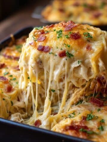 Homemade chicken spaghetti casserole in a black baking dish, topped with golden melted cheese, crispy bacon, and parsley, with a gooey cheese pull revealing creamy spaghetti and shredded chicken inside.