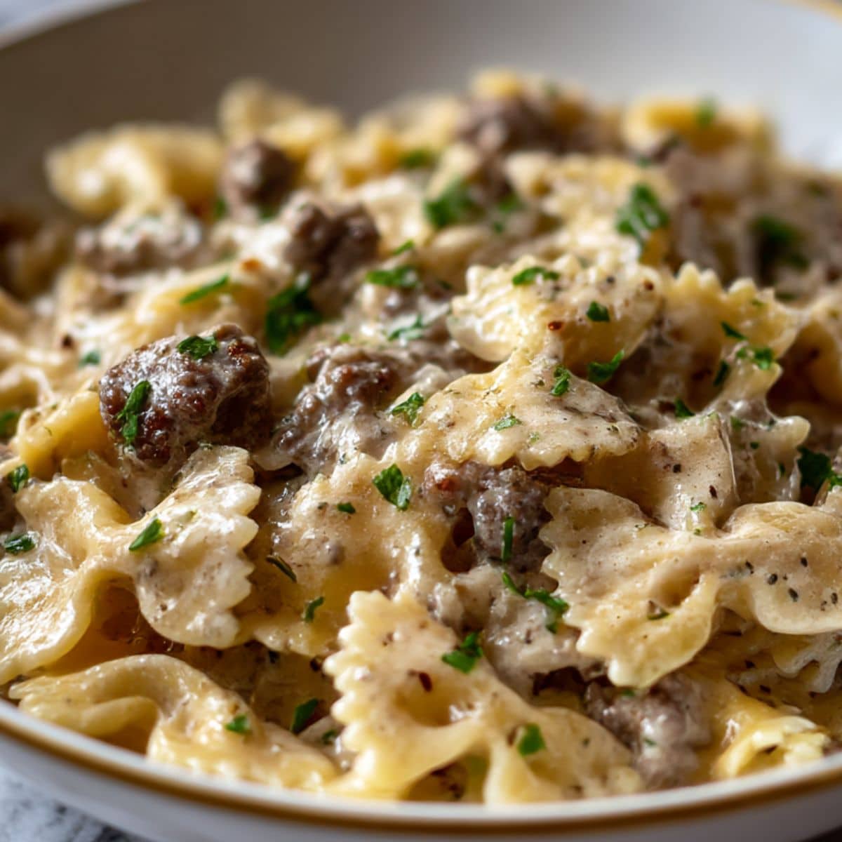 Creamy beef and bowtie pasta coated in rich Alfredo sauce, topped with fresh parsley and served in a white bowl on a marble countertop.