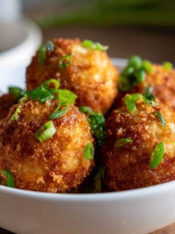 Fried mashed potato bites with golden brown crust, topped with chopped green onions, served in a white bowl