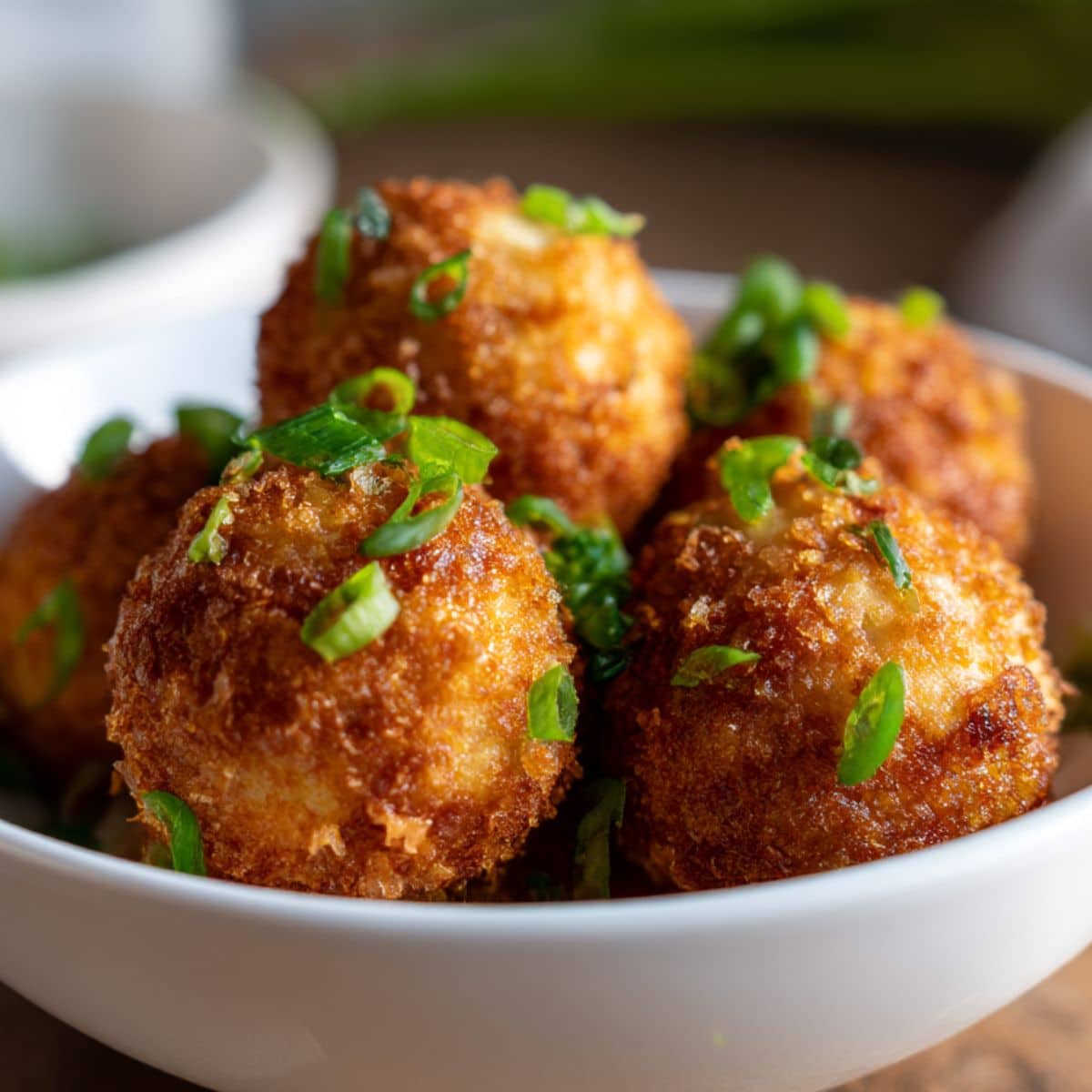 Fried mashed potato bites with golden brown crust, topped with chopped green onions, served in a white bowl
