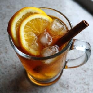 Homemade orange cinnamon tea in clear glass with ice cubes, orange wheel garnish and cinnamon stick.