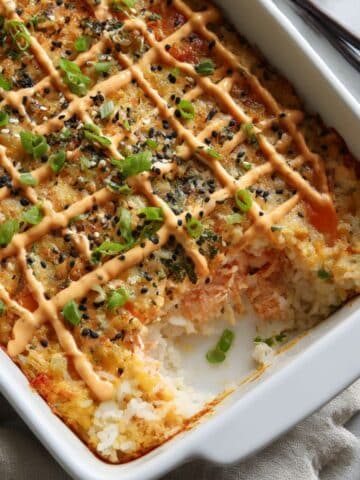 Homemade salmon sushi bake topped with spicy mayo, sesame seeds, and green onions, with a corner scooped out to reveal creamy salmon and rice layers.