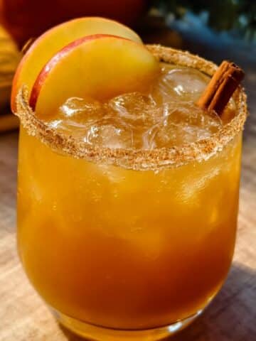 Apple cider margarita with cinnamon sugar rim, apple slices, and cinnamon stick served over ice in a glass.