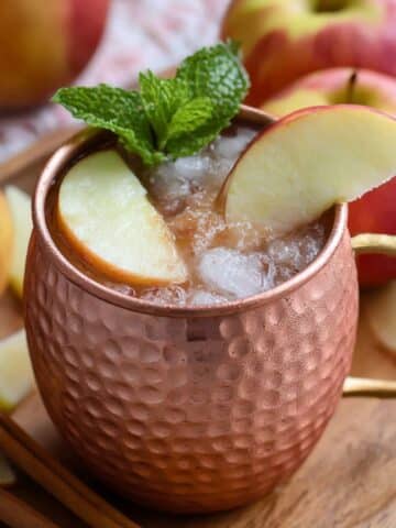 Apple cider Moscow Mule served in a chilled copper mug with crushed ice, apple slices, mint, and cinnamon sticks.
