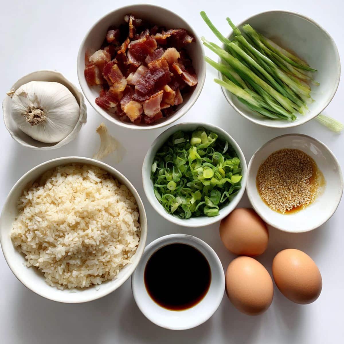 Ingredients for homemade breakfast fried rice arranged on a white counter, including boowls of rice, bacon, eggs, and sauces.