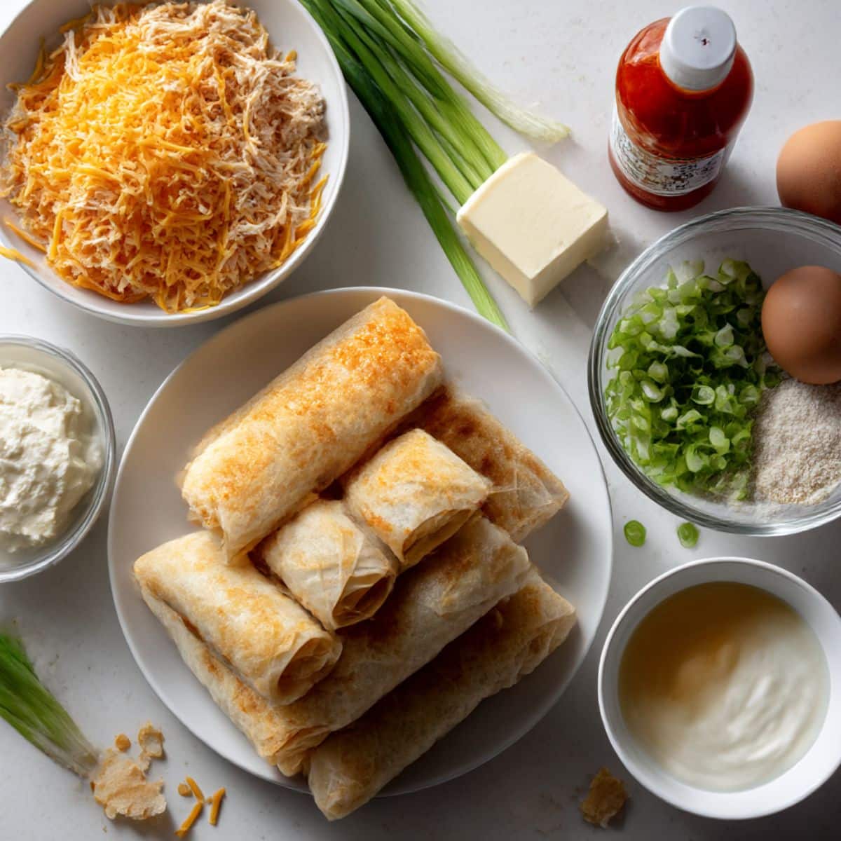 Overhead shot of buffalo chicken dip egg roll ingredients on a white kitchen counter including chicken, cream cheese, buffalo sauce, cheddar, wrappers, and ranch.