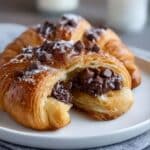 Flaky, golden-brown chocolate croissant filled with melted chocolate chips and topped with powdered sugar, served on a white plate for a mouthwatering pastry presentation.