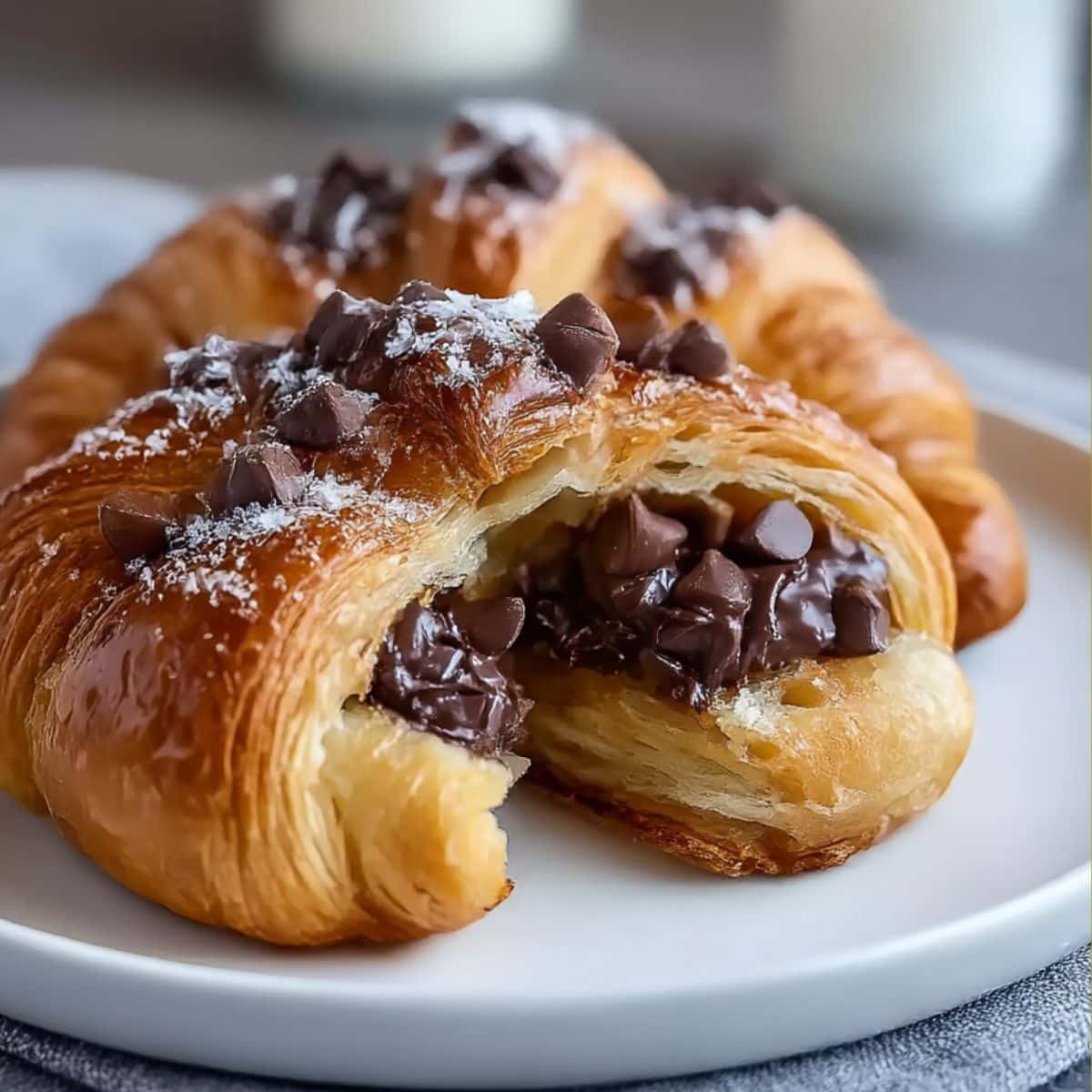 Flaky, golden-brown chocolate croissant filled with melted chocolate chips and topped with powdered sugar, served on a white plate for a mouthwatering pastry presentation.