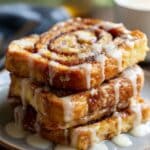 Caramelized cinnamon roll French toast slices stacked on a plate, swirled with cinnamon sugar and topped with icing glaze, golden-brown edges with a sticky, juicy finish.