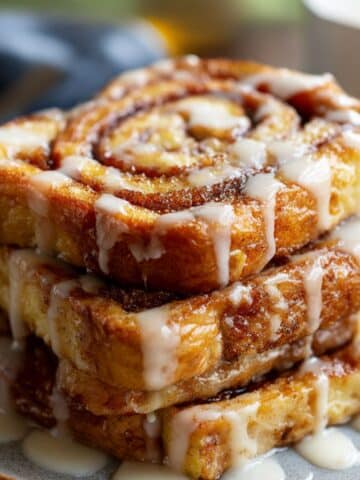 Caramelized cinnamon roll French toast slices stacked on a plate, swirled with cinnamon sugar and topped with icing glaze, golden-brown edges with a sticky, juicy finish.