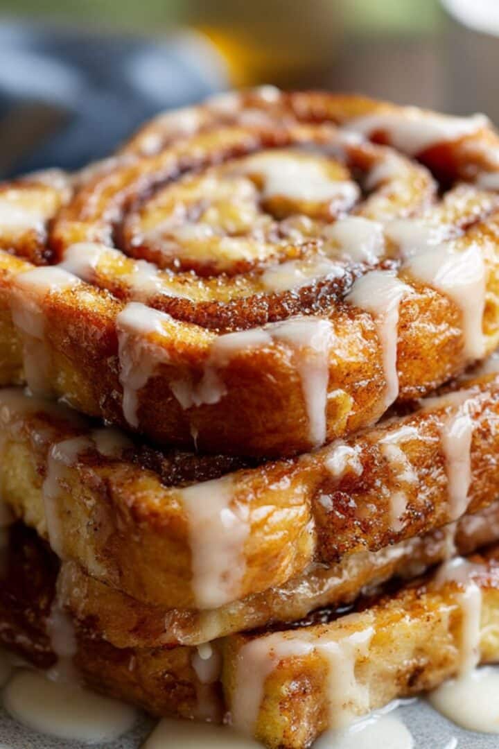 Caramelized cinnamon roll French toast slices stacked on a plate, swirled with cinnamon sugar and topped with icing glaze, golden-brown edges with a sticky, juicy finish.