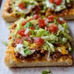 lose-up of homemade crescent roll taco pizza topped with ground beef, melted cheese, lettuce, tomatoes, and sour cream on golden flaky crust. Perfect easy weeknight dinner idea.