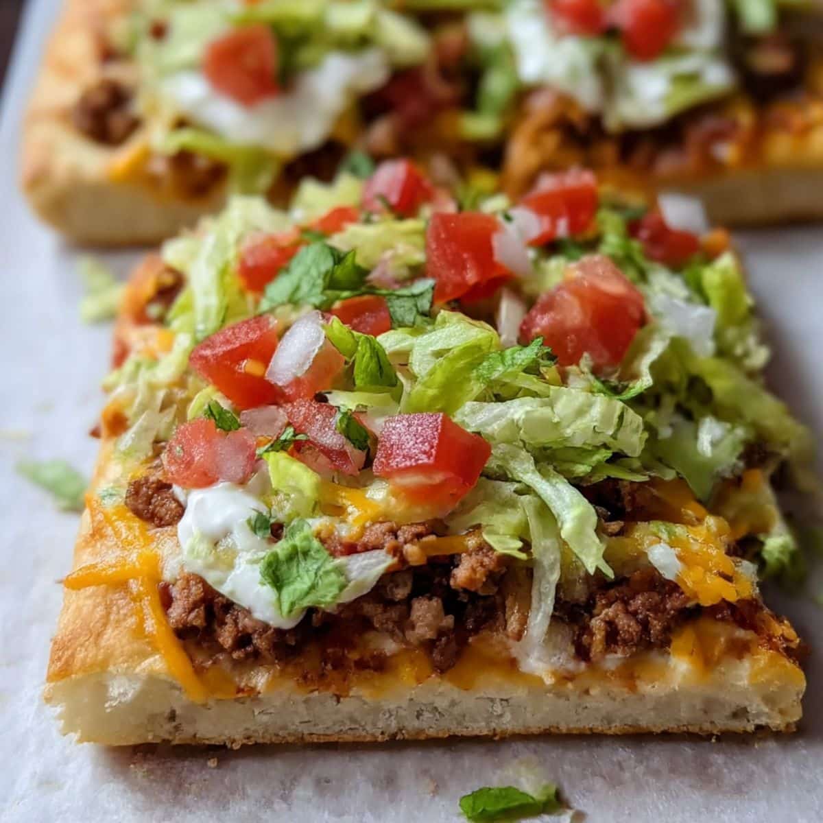 lose-up of homemade crescent roll taco pizza topped with ground beef, melted cheese, lettuce, tomatoes, and sour cream on golden flaky crust. Perfect easy weeknight dinner idea.