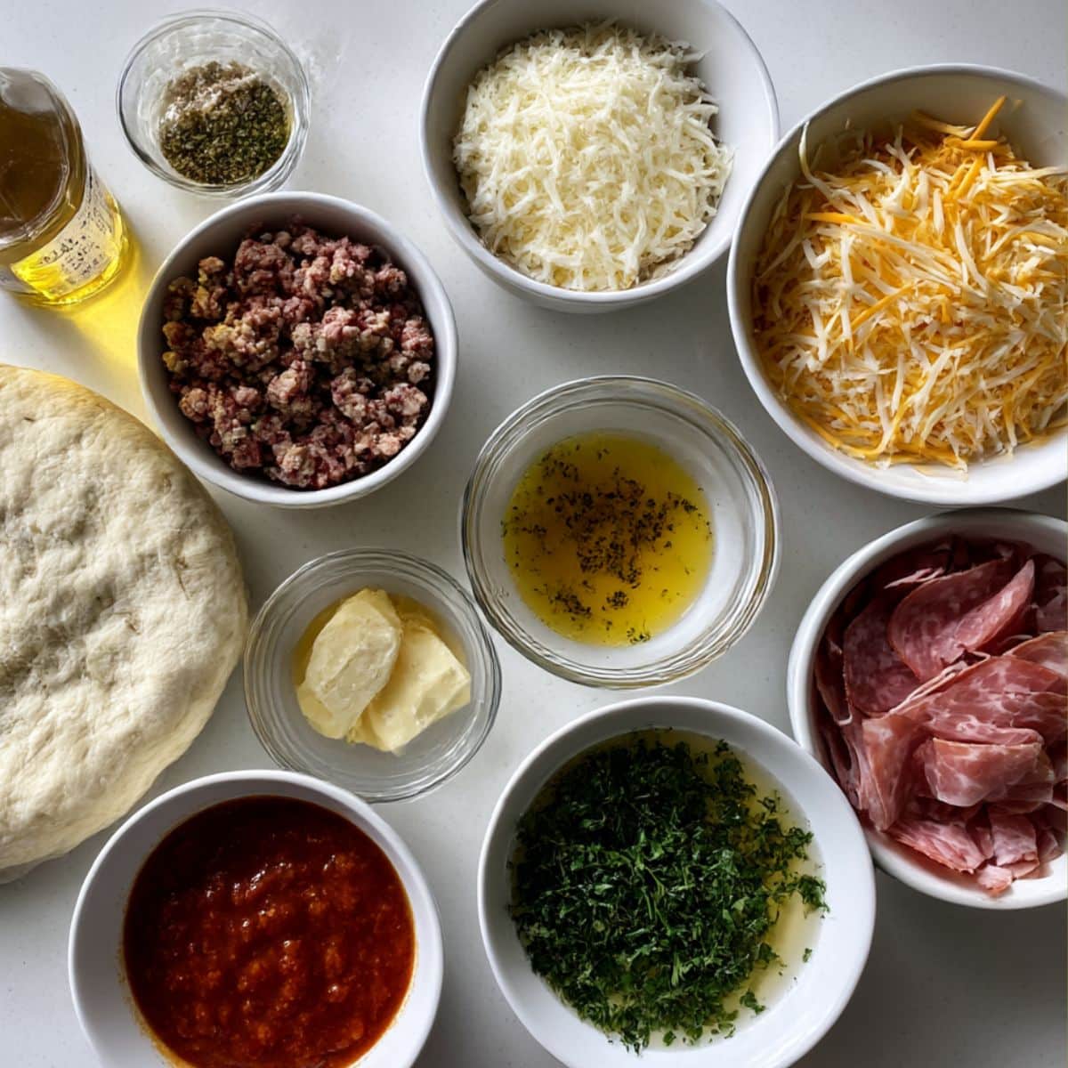 Overhead photo of pizza dough, meats, cheeses, sauce, and garlic butter ingredients for homemade garbage bread.