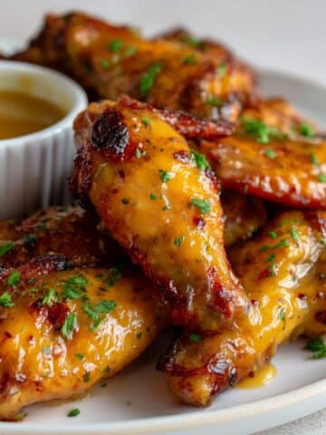 Sticky honey mustard chicken wings coated in a thick, glossy glaze, garnished with fresh chopped parsley and served on a white plate with a ramekin of dipping sauce in the background.