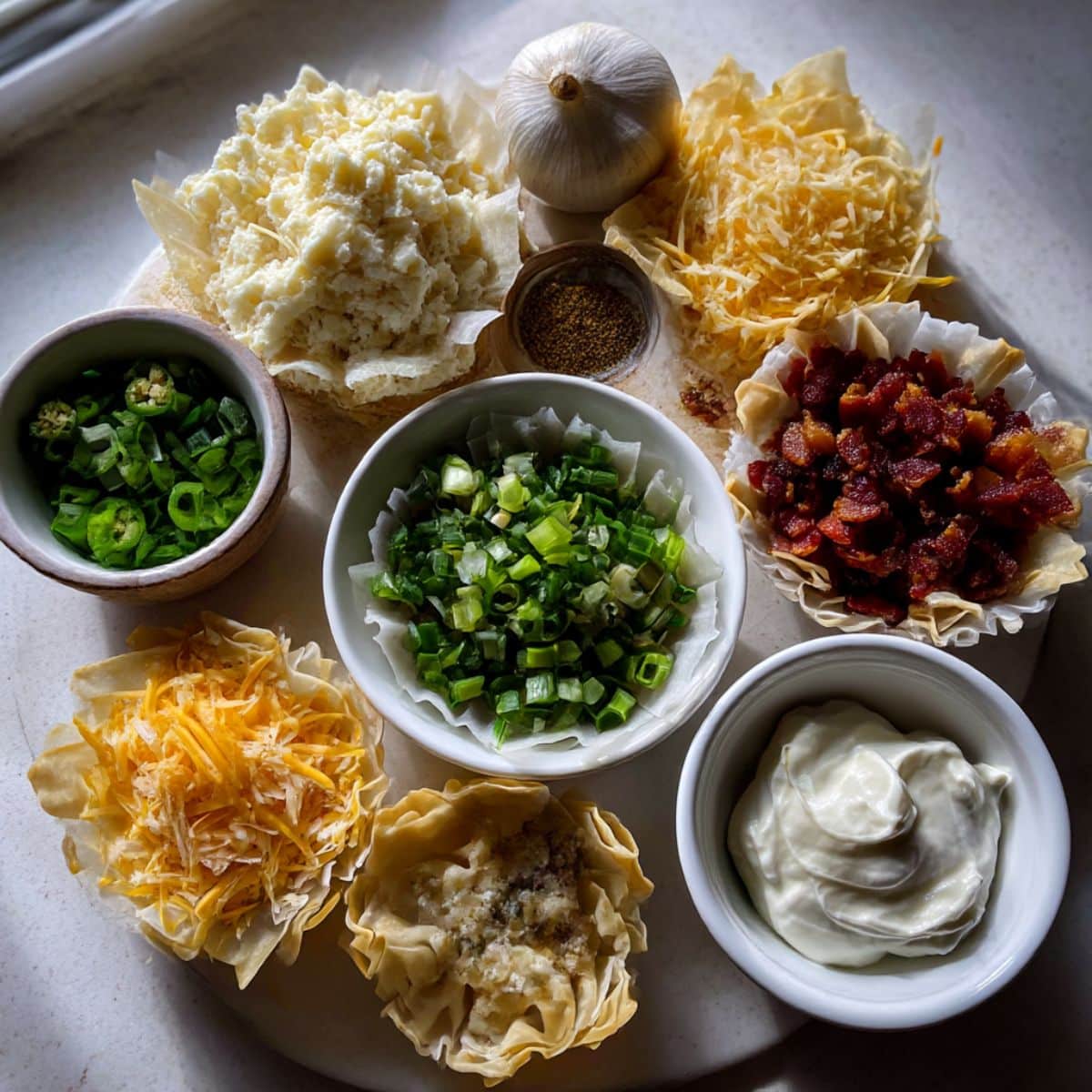 Overhead view of ingredients for jalapeño popper wonton cups including wonton wrappers, cream cheese, cheddar, bacon, jalapeños, and green onions on a white kitchen counter.