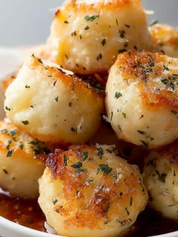 Golden parmesan bread bites brushed with garlic butter and sprinkled with herbs, served warm in a white bowl.