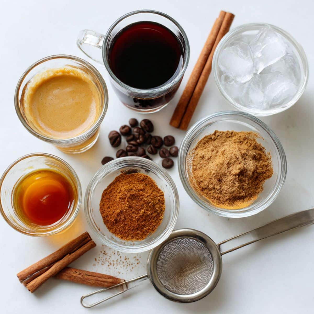All ingredients for homemade pumpkin spice espresso martini on a white counter, including espresso, pumpkin spice syrup, vodka, coffee liqueur, cinnamon, and ice.