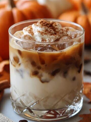 Pumpkin spice white russian cocktail over ice topped with whipped cream and cinnamon, surrounded by pumpkins and fall leaves.