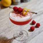 Raspberry gin sour cocktail in a coupe glass with frothy foam, garnished with raspberries and lemon twist on a marble counter.