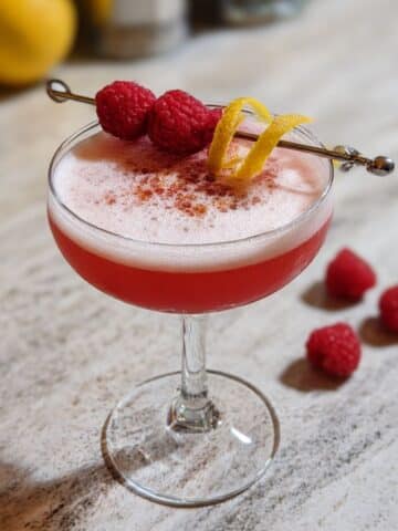 Raspberry gin sour cocktail in a coupe glass with frothy foam, garnished with raspberries and lemon twist on a marble counter.