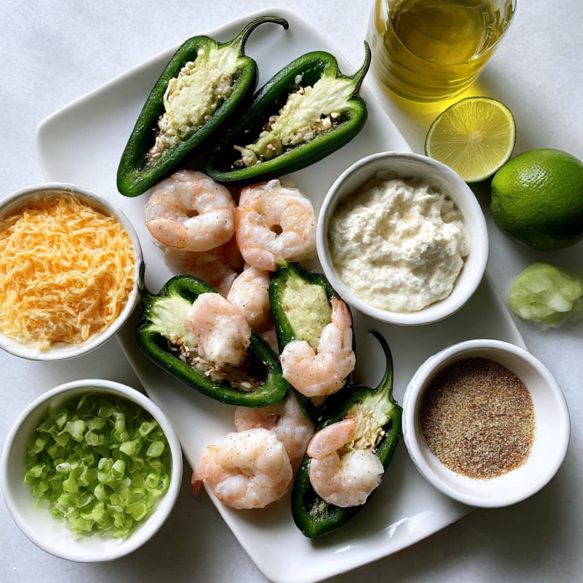 Overhead view of ingredients for shrimp stuffed jalapeño peppers including fresh jalapeños, shrimp, cream cheese, cheddar, garlic, and lime on a white kitchen counter.