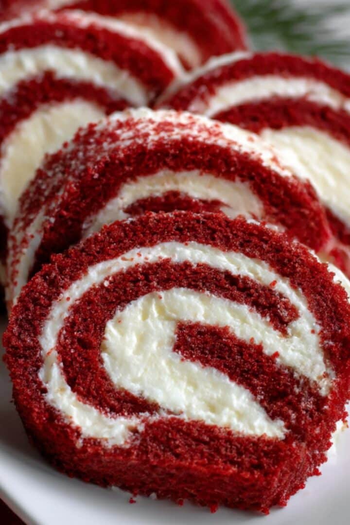 A festive Christmas red velvet cake roll with creamy white filling, sliced and served on a white plate; perfect for Christmas dessert tables.